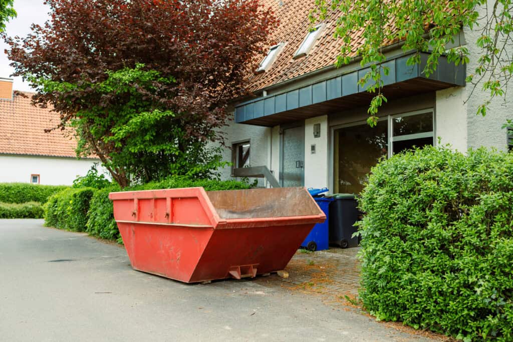 Large red dumpster outside residential building with greenery.