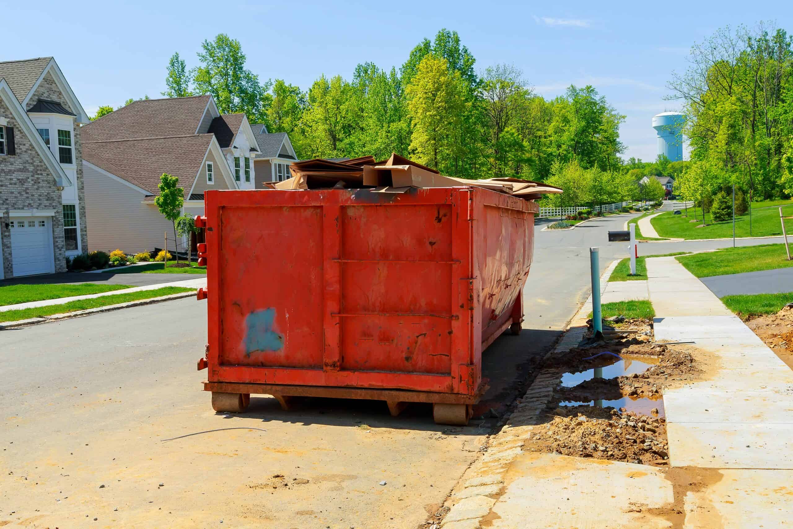 Red dumpster on residential street with green trees and houses in background.